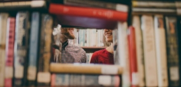 Fotografía de una pila de libros a través de la cual se ve parte de la cara de dos estudiantes