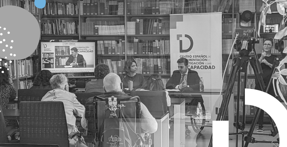 Fotografía de la charla en la sala del CEDID. Se ve a Carmen Vázquez junto a José Antonio Rueda Monroy tras una mesa y a la intérprete de lengua de signos. Frente a ellos el público asistente.